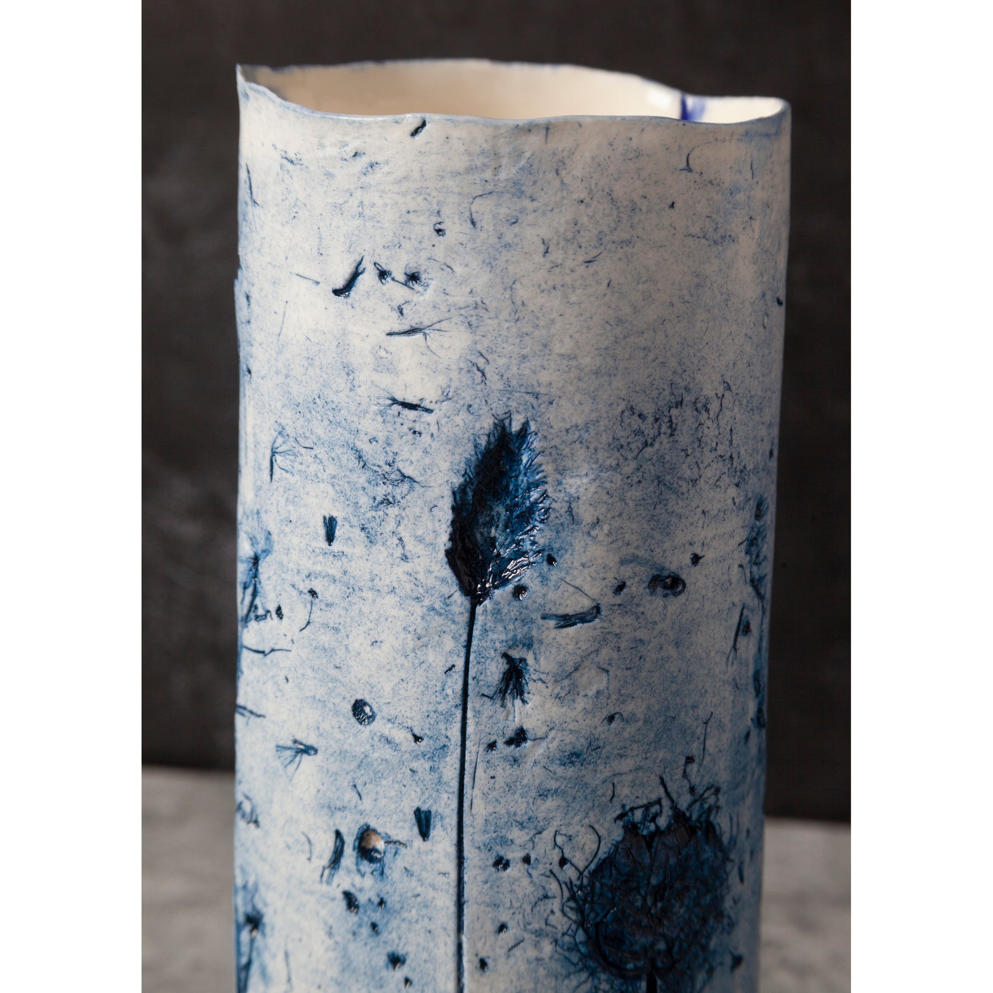 Cylindrical ceramic vase with textured surface and blue feather-like design on a dark background